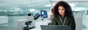 A woman works intently on a computer at a desk in a lab with a microscope next to her